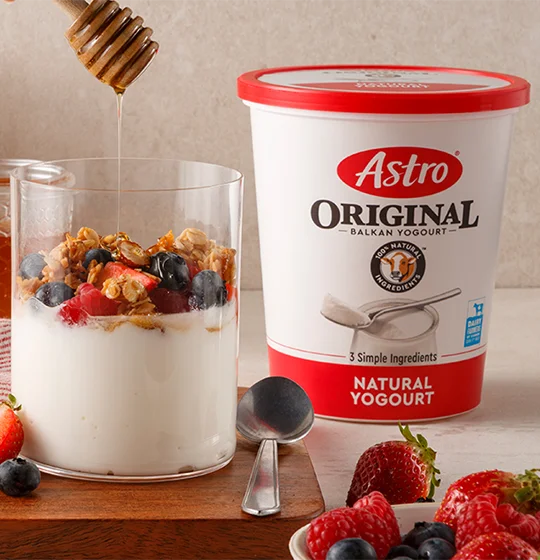 Astro Plain 6% yogurt tub on a table with yogurt in a glass with honey, fruit and grains